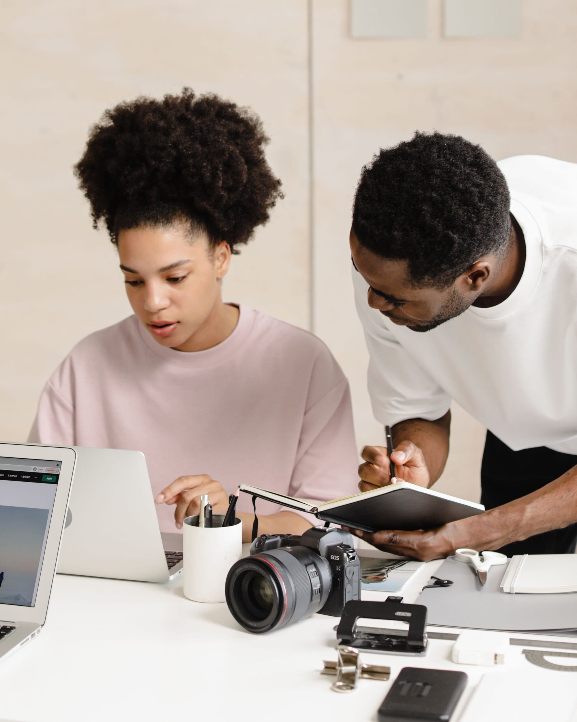 Two people at a bright white desk with a laptop, a camera, a tablet and printed sheets, one making notes
