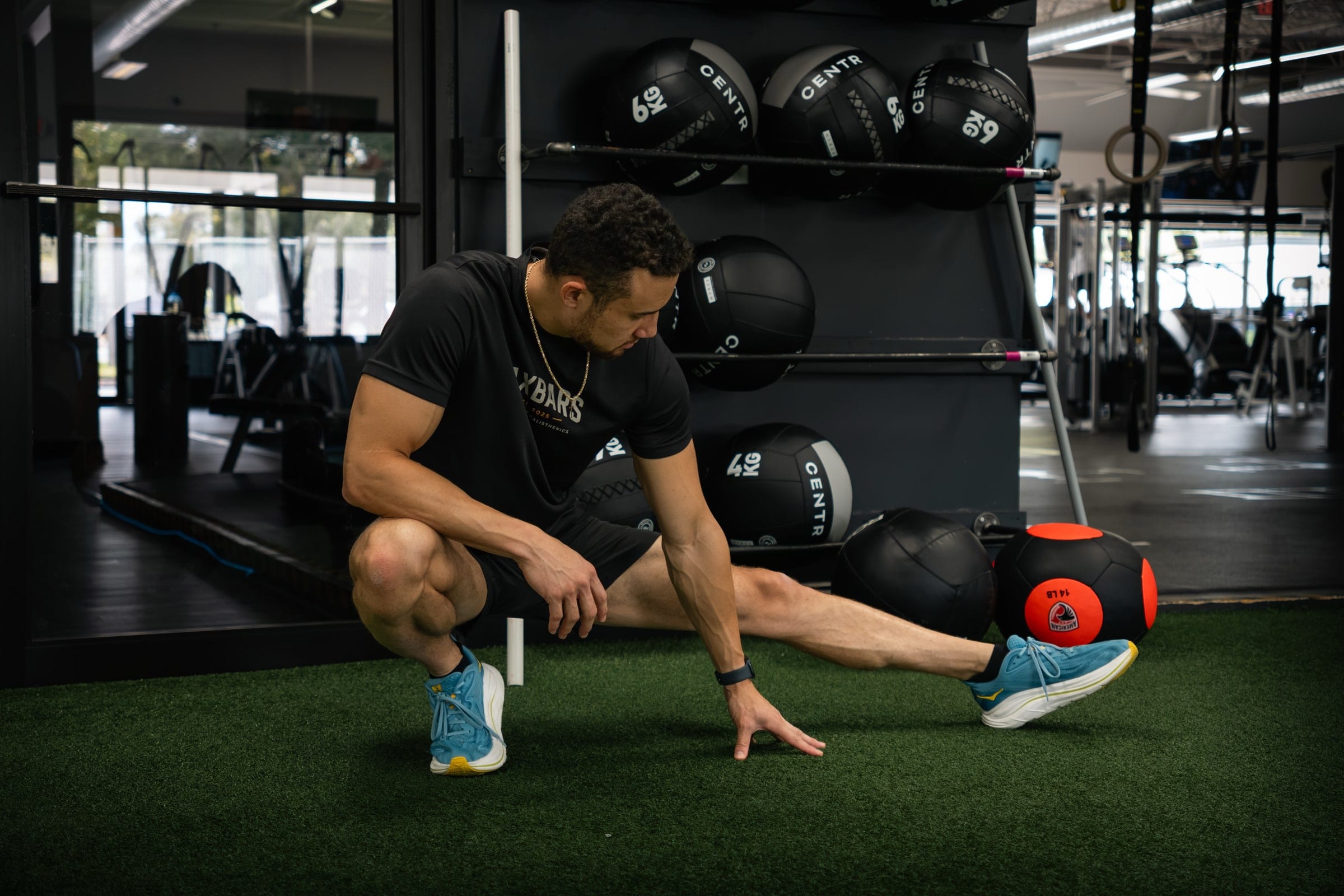 An athlete in black crouches into a mobility stretch on gym turf in front of a rack of medicine balls