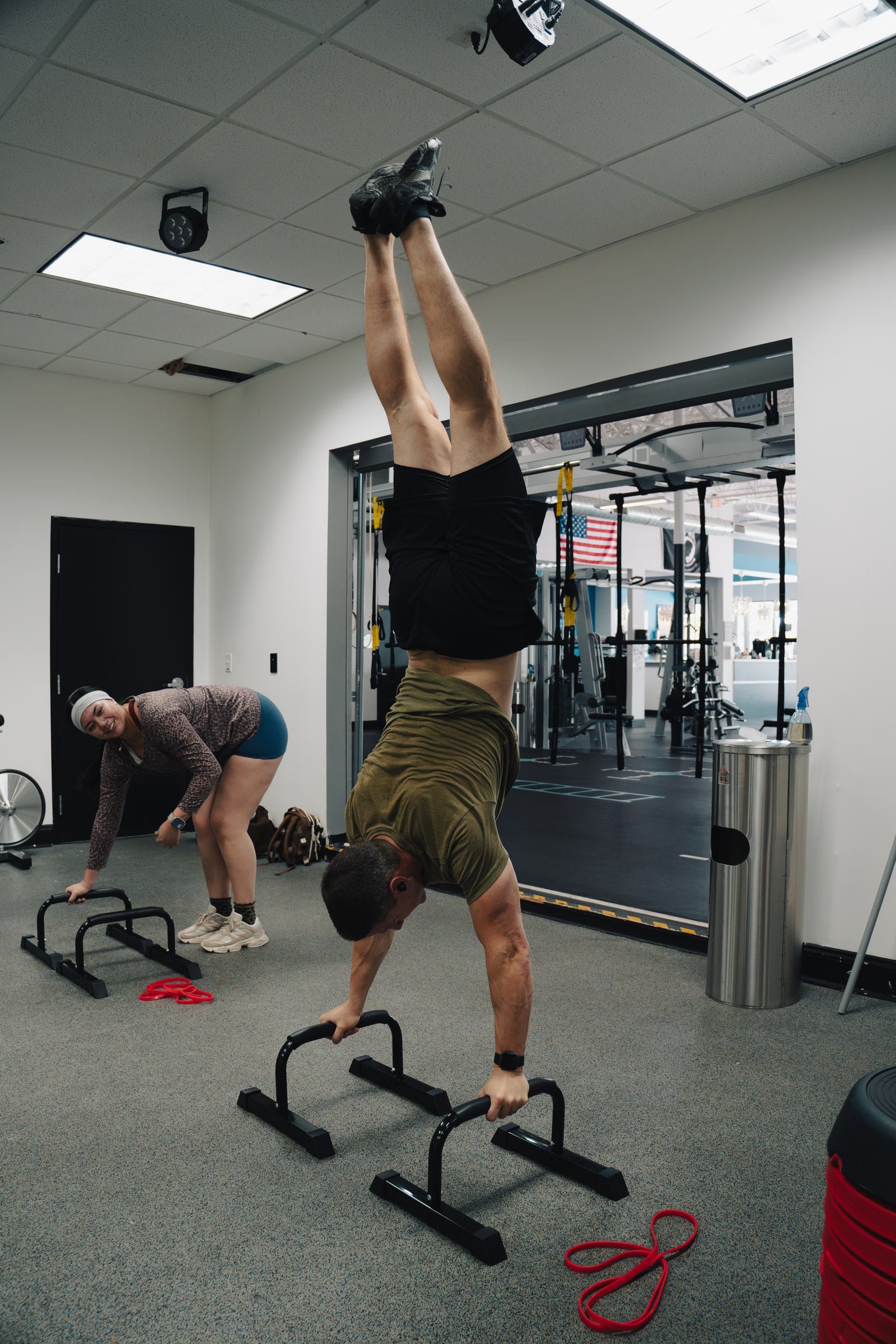 A participant holds a handstand on parallettes while another stretches behind him during a calisthenics class
