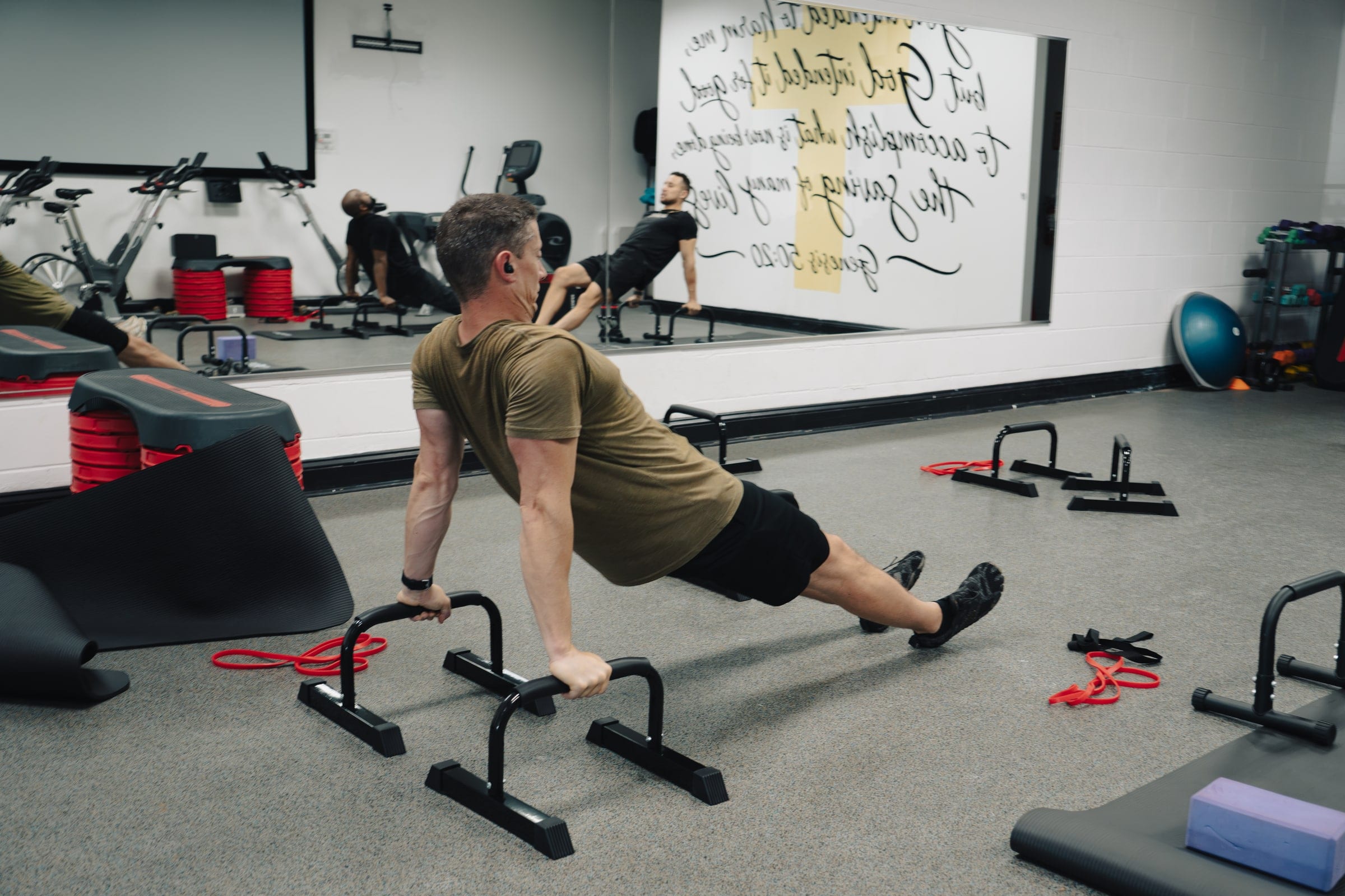 A participant holds a strength move on parallettes during a calisthenics class, the gym mirrored behind him