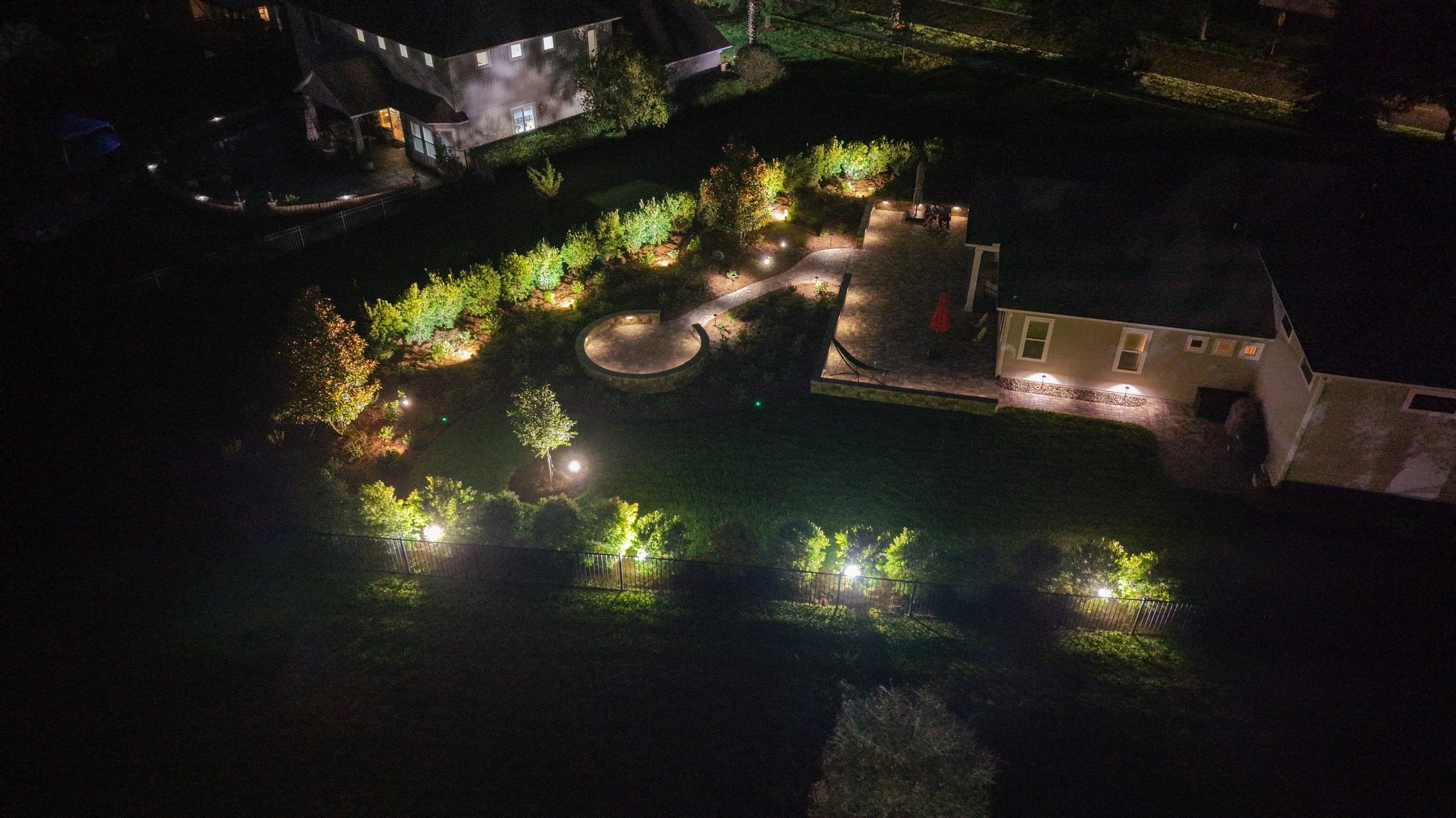 Night aerial view of a backyard with lighting along the garden beds, a lit patio and the house beside it
