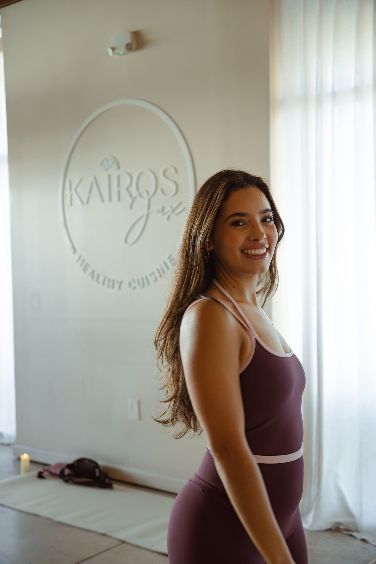 A woman in plum activewear smiles over her shoulder beside the Kairos Healthy Cuisine logo on a white wall