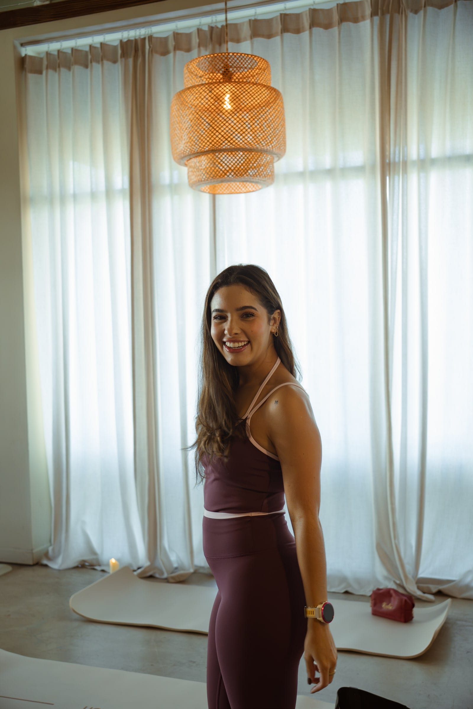 A woman in plum activewear smiles by sheer curtains under a rattan pendant in soft window light
