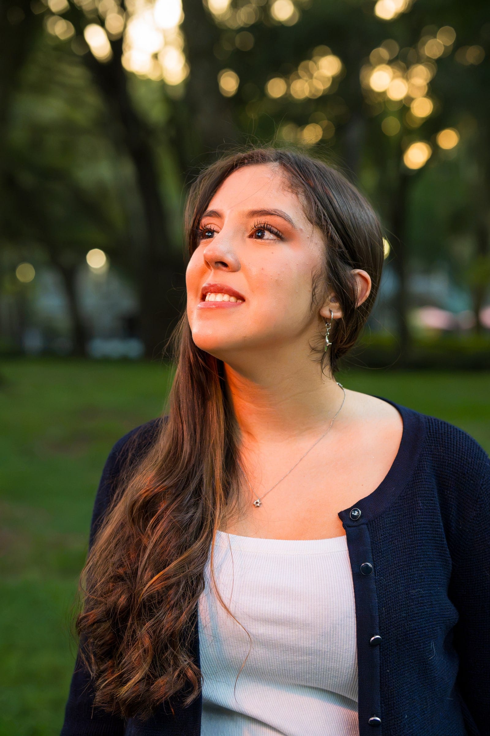 A smiling portrait subject in a navy cardigan looks past the camera in golden-hour light, park trees glowing behind her