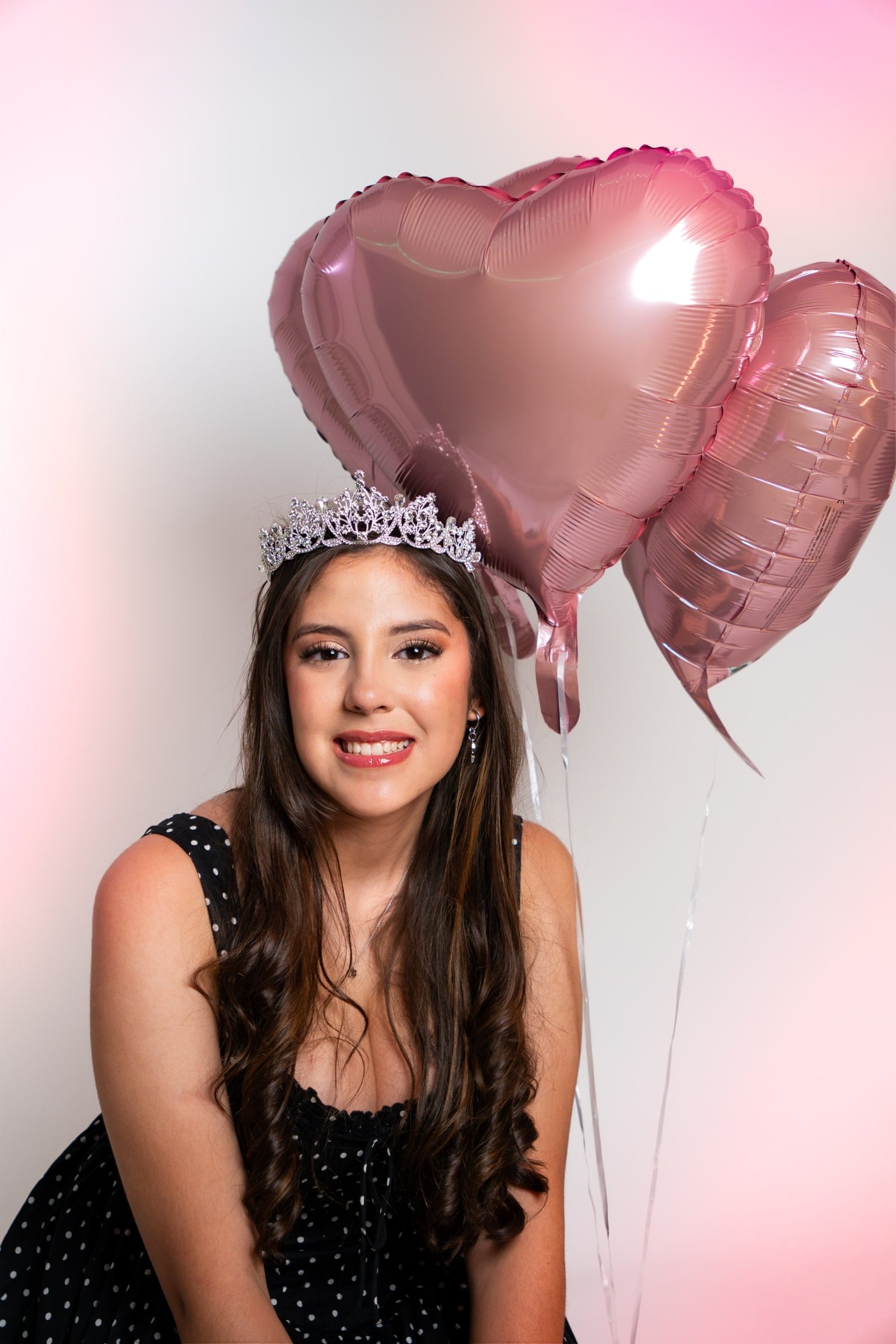 Studio celebration portrait: a smiling subject in a tiara and polka-dot dress beside pink heart balloons