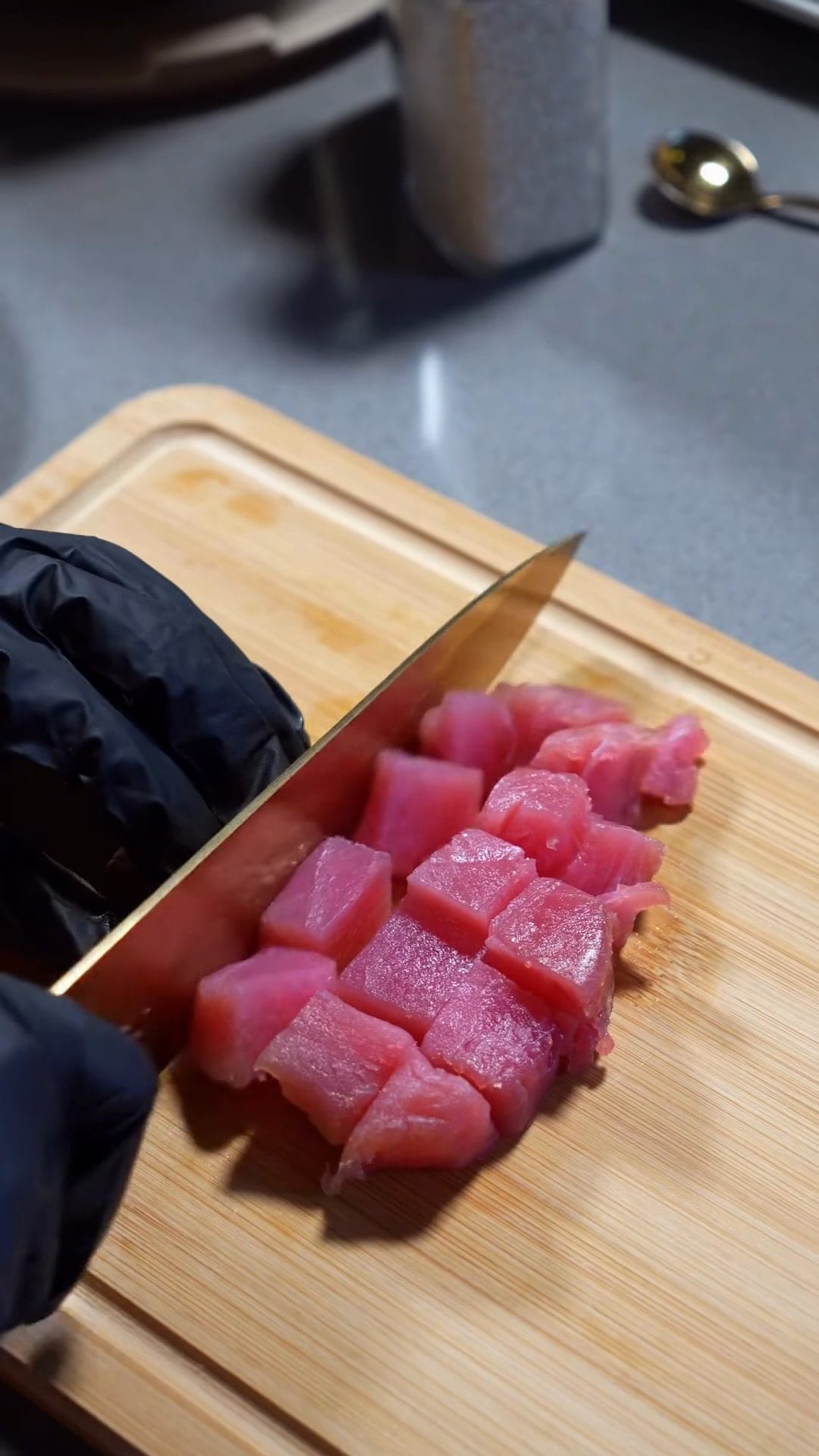 Frame from a recipe film: a chef's knife sliding through cubes of fresh tuna on a bamboo board
